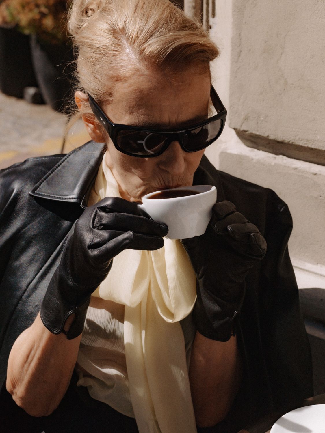 A woman in sunglasses and a leather coat drinking coffee outside, ready for a press day